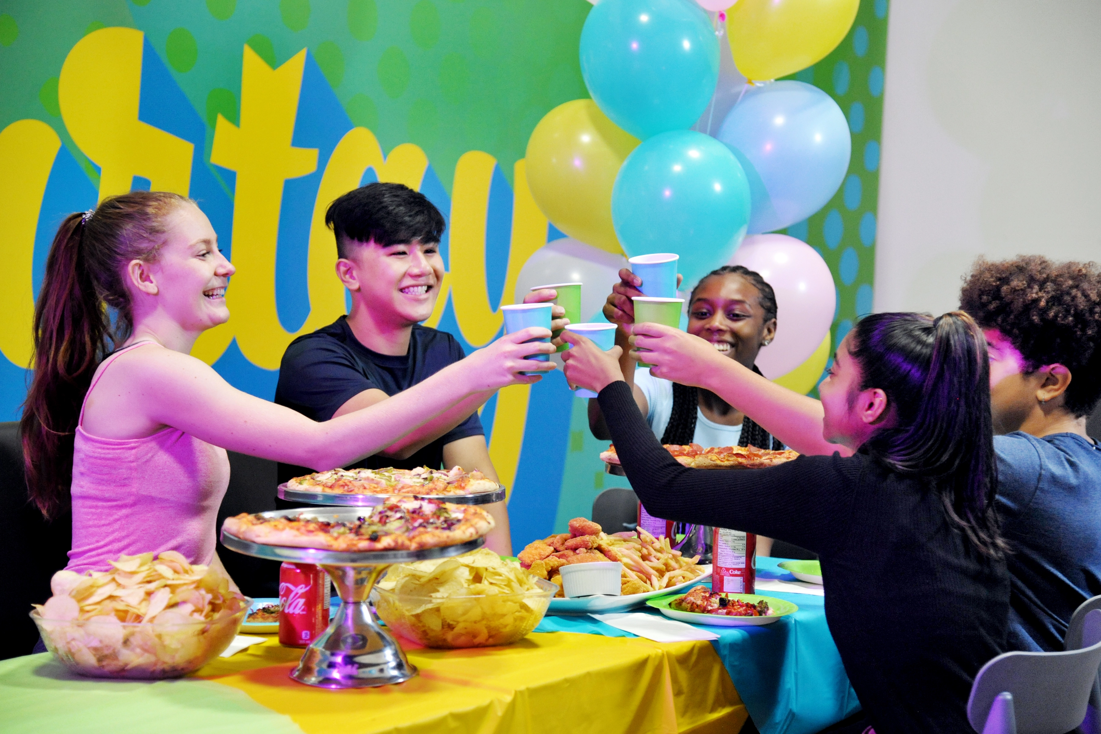 Adults eating pizza and  holding up their drinks at a party with their colleagues.
